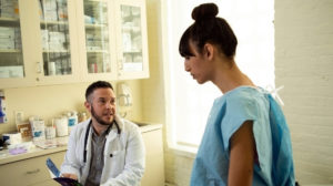 A transgender woman in a hospital gown speaking to her doctor, a transgender man, in an exam room.