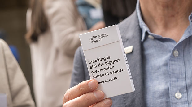 A photograph of someone holding a cigarette packet that reads 'Smoking is still the biggest preventable cause of cancer'.