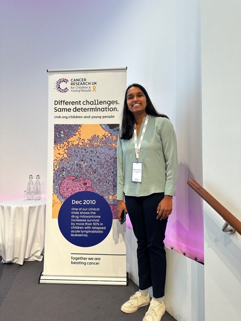 Shaumya standing in front of a poster for Cancer Research UK's Children's and Young People's Conference, reading 'Different challenges, same determination.'