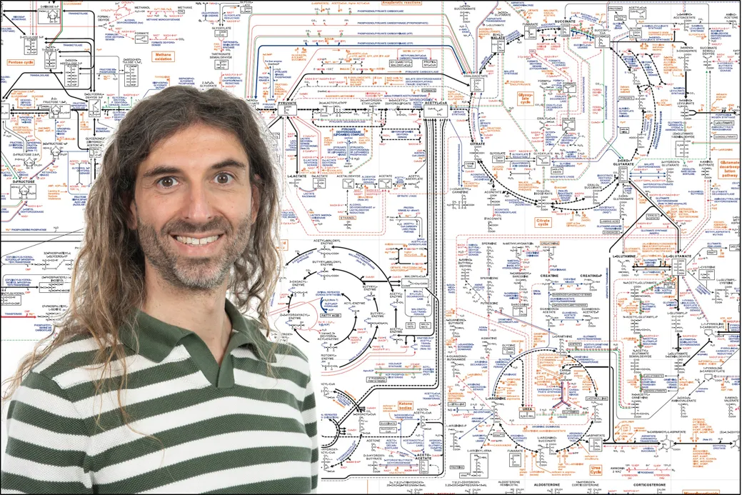 Dr Saverio Tardito smiling in front of a board detailing some of the metabolic pathways this research looks at.