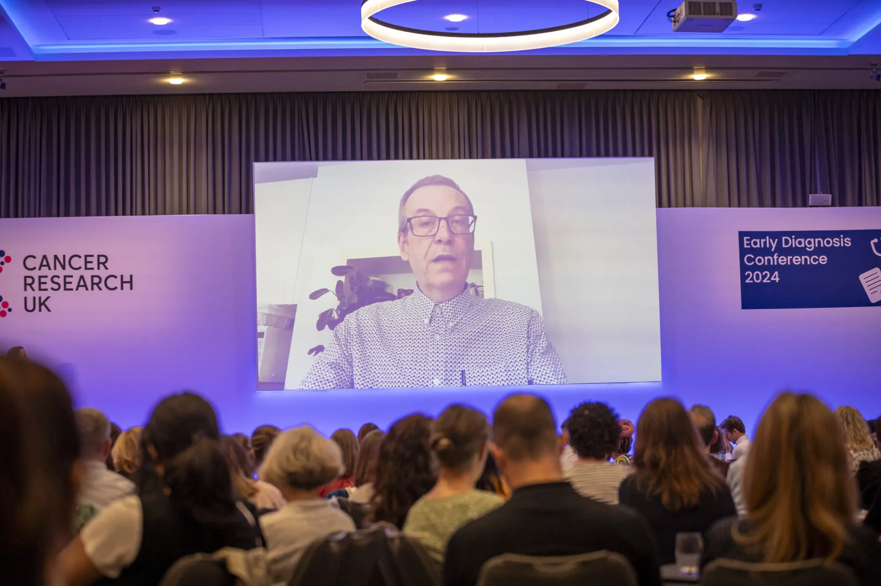 Michael, on a large screen, speaking at Cancer Research UK's Early Diagnosis conference