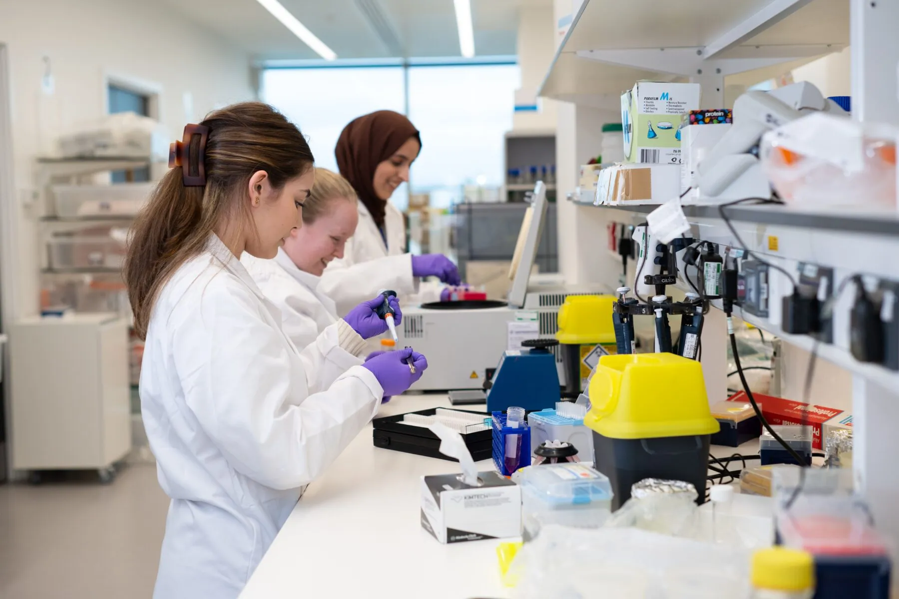 A group of three female Cancer Research UK scientists working with samples at a lab bench. They are smiling, talking and wearing lab coats.