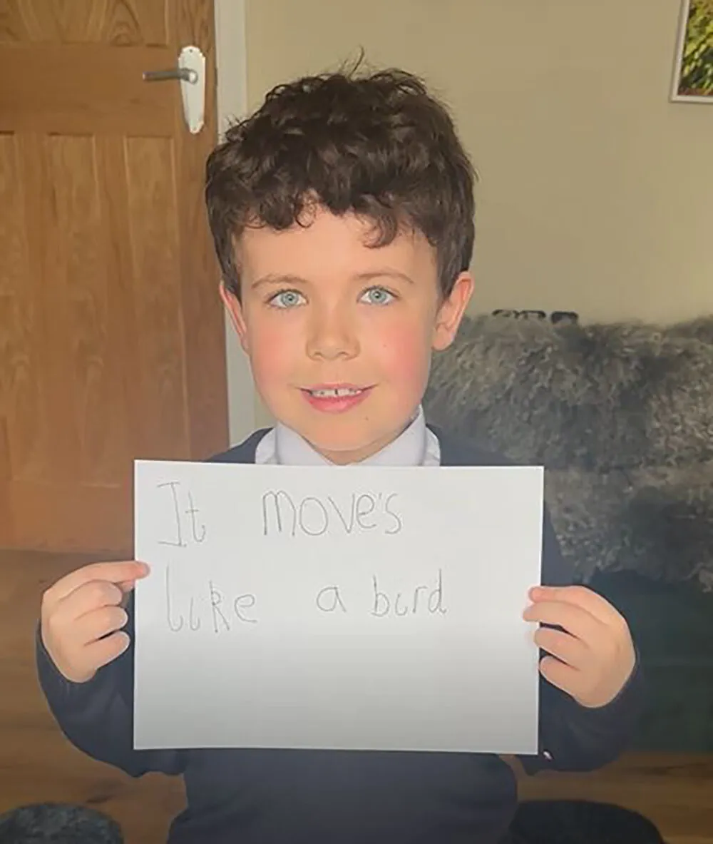 Benny, a young boy with dark hair, stands holding a sign that says 'moves like a bird'