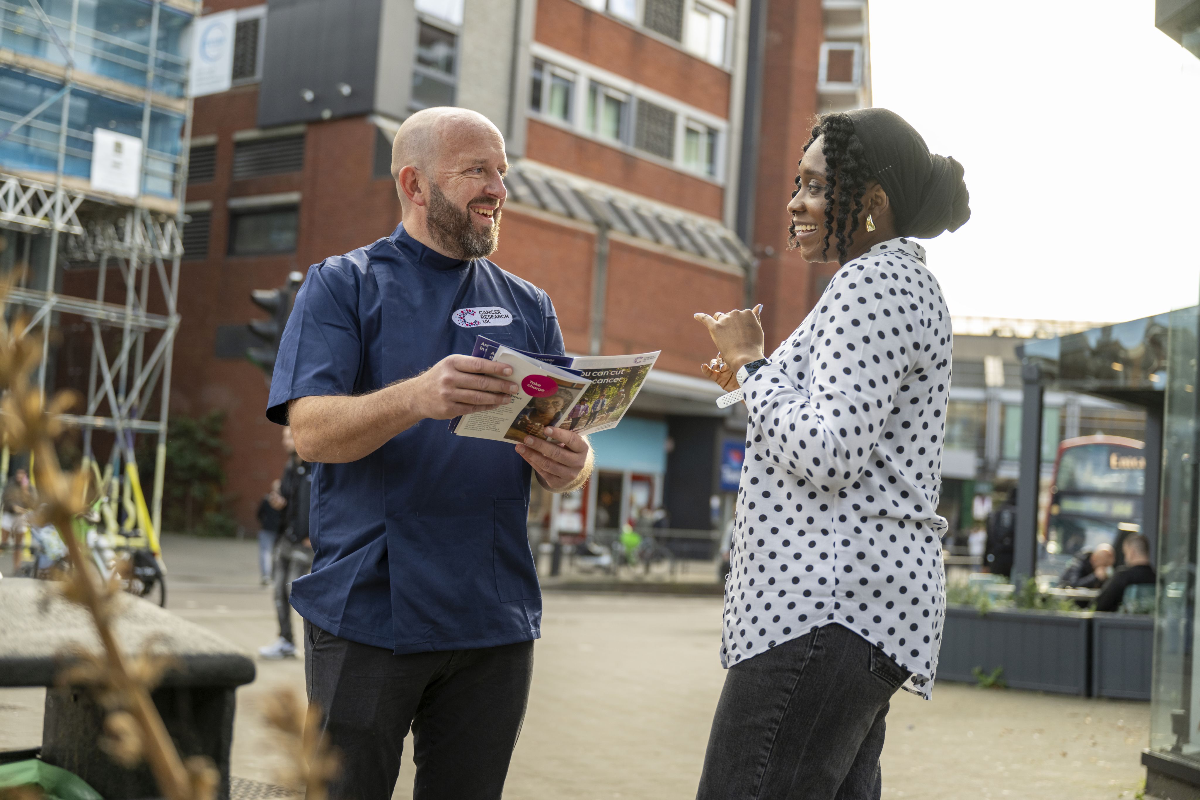 A male Cancer Research UK nurse who is holding leaflets speaks to a woman