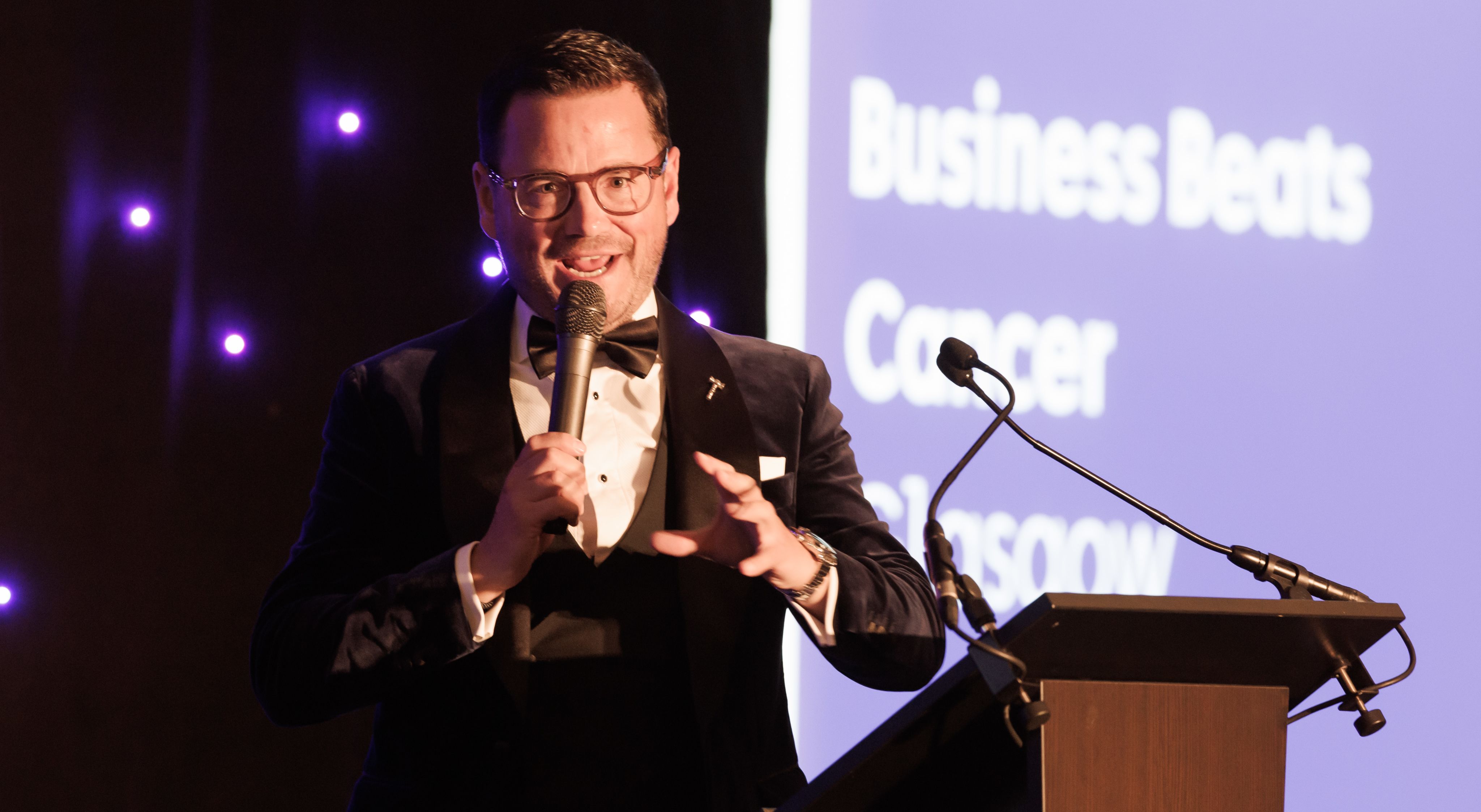A man with a microphone speaks at the Glasgow Business Beats Cancer event