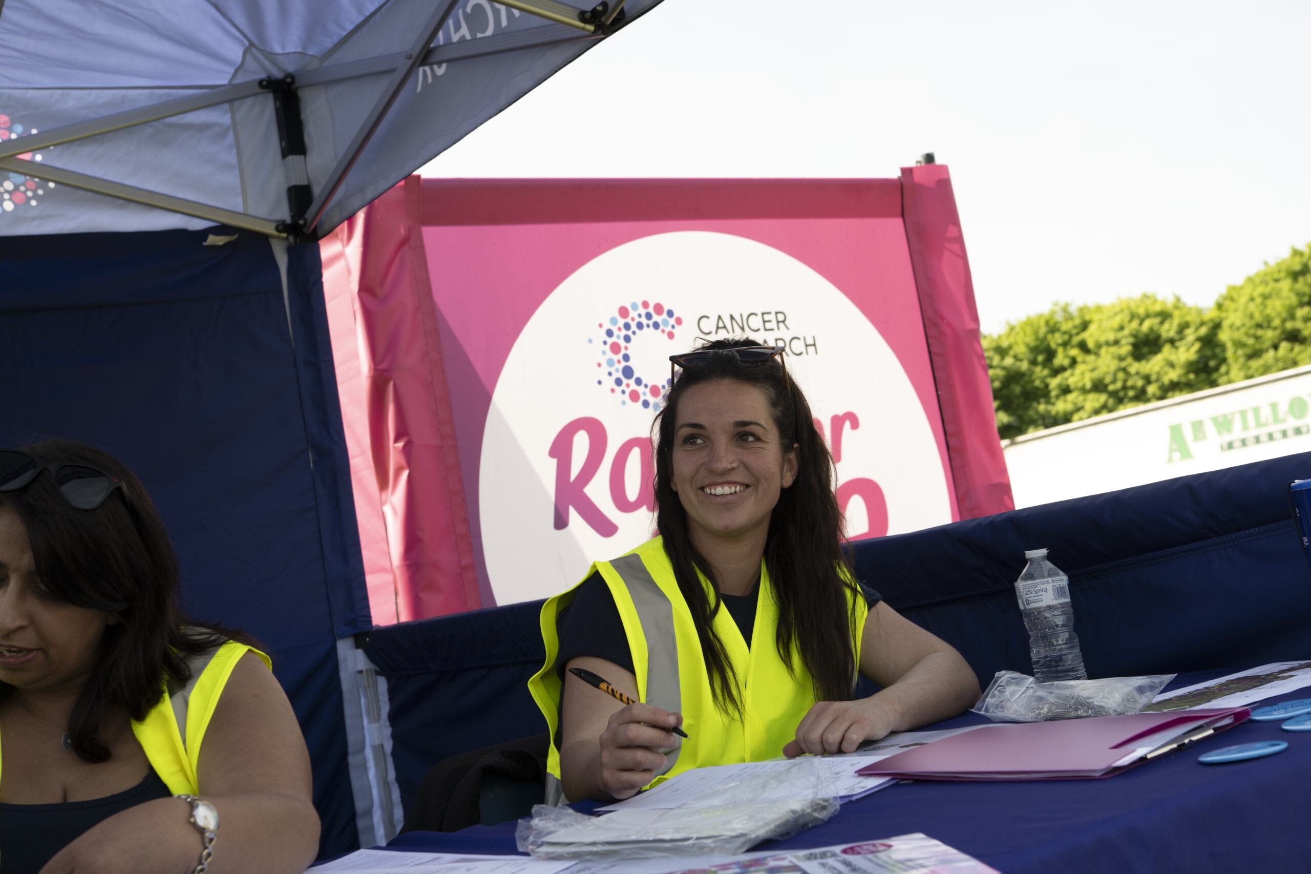 A female volunteer with dark hair and a high vis vest sits in front of a race for life sign