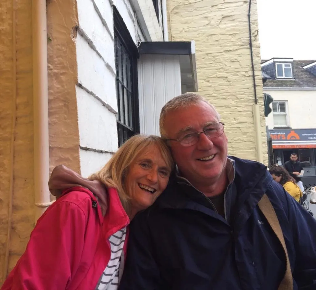 David together with his wife Marion sat outside a cafe. Marion is leaning on David's shoulder, and David's head is resting against hers. They're both smiling at the camera.