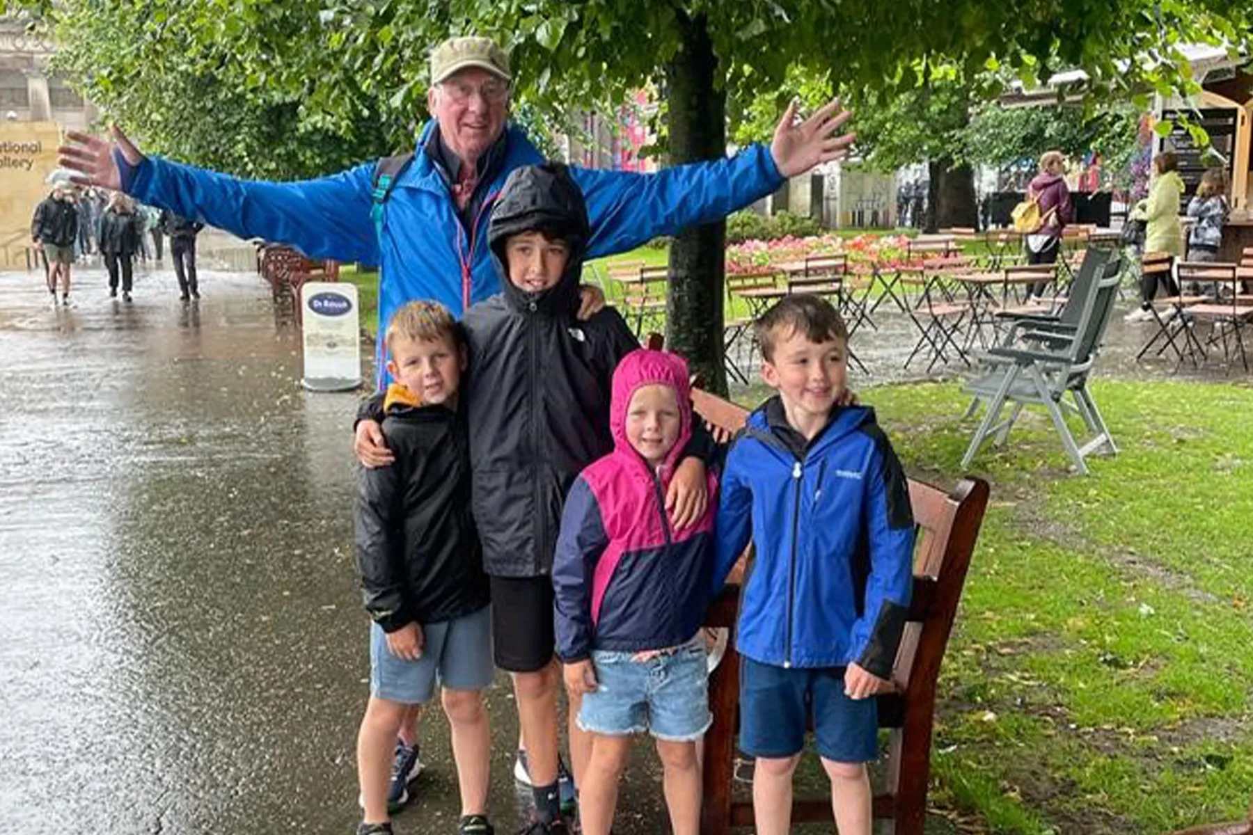 David on a day out with his four grandchildren. They are stood together under a tree on a rainy summer day, dressed in raincoats and shorts. The children have their arms round each other. David is behind them with his arms spread wide.