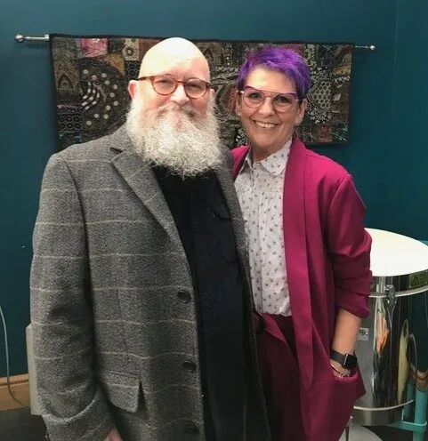 Sue and her husband George dressed in suits. George's jacket has a grey check pattern. Sue is wearing a soft cut blazer in magenta. George has a large white beard and Sue has short purple hair. Both are wearing glasses that match their outfits.