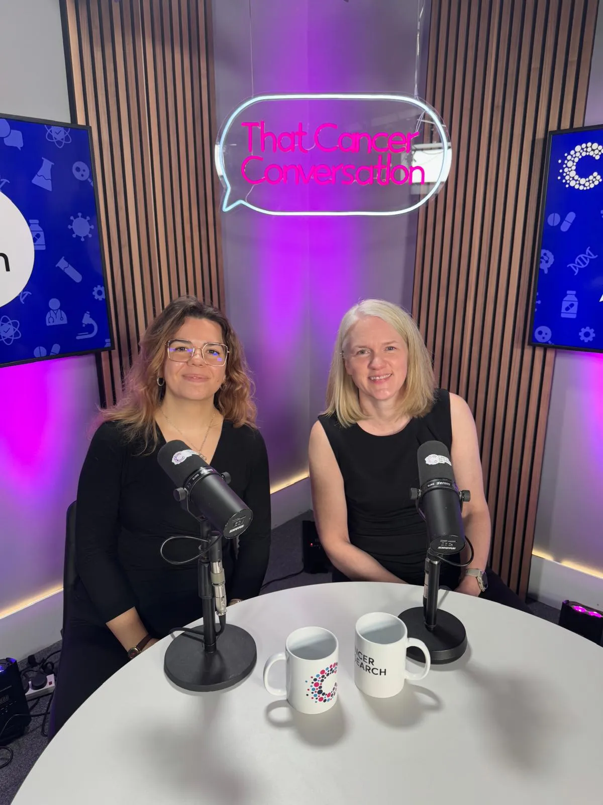 Sophie and Heather in front of podcast microphones smiling to the camera