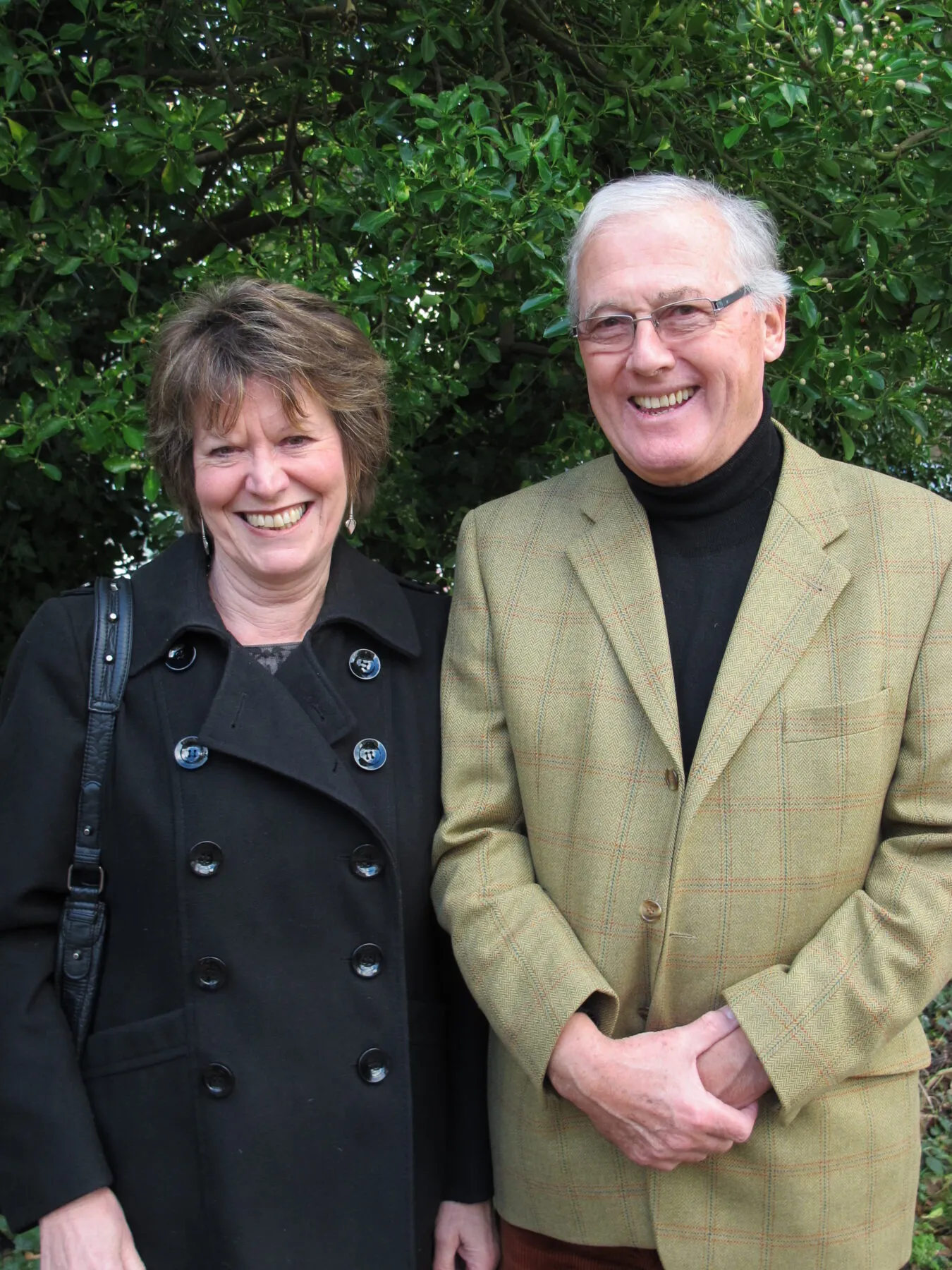 Peter and his wide Ali standing smiling in front of some trees. Ali is wearing a black trenchcoat and Peter is wearing a tweed jacket.