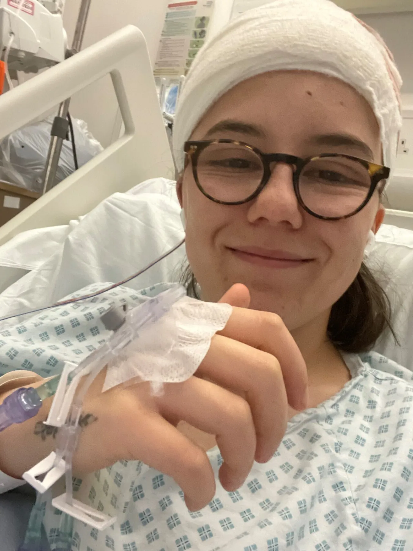 A selfie of Jessie in her hospital bed after her brain surgery. . She is smiling and giving a thumbs up to the camera. She is wearing glasses and her head is bandaged.