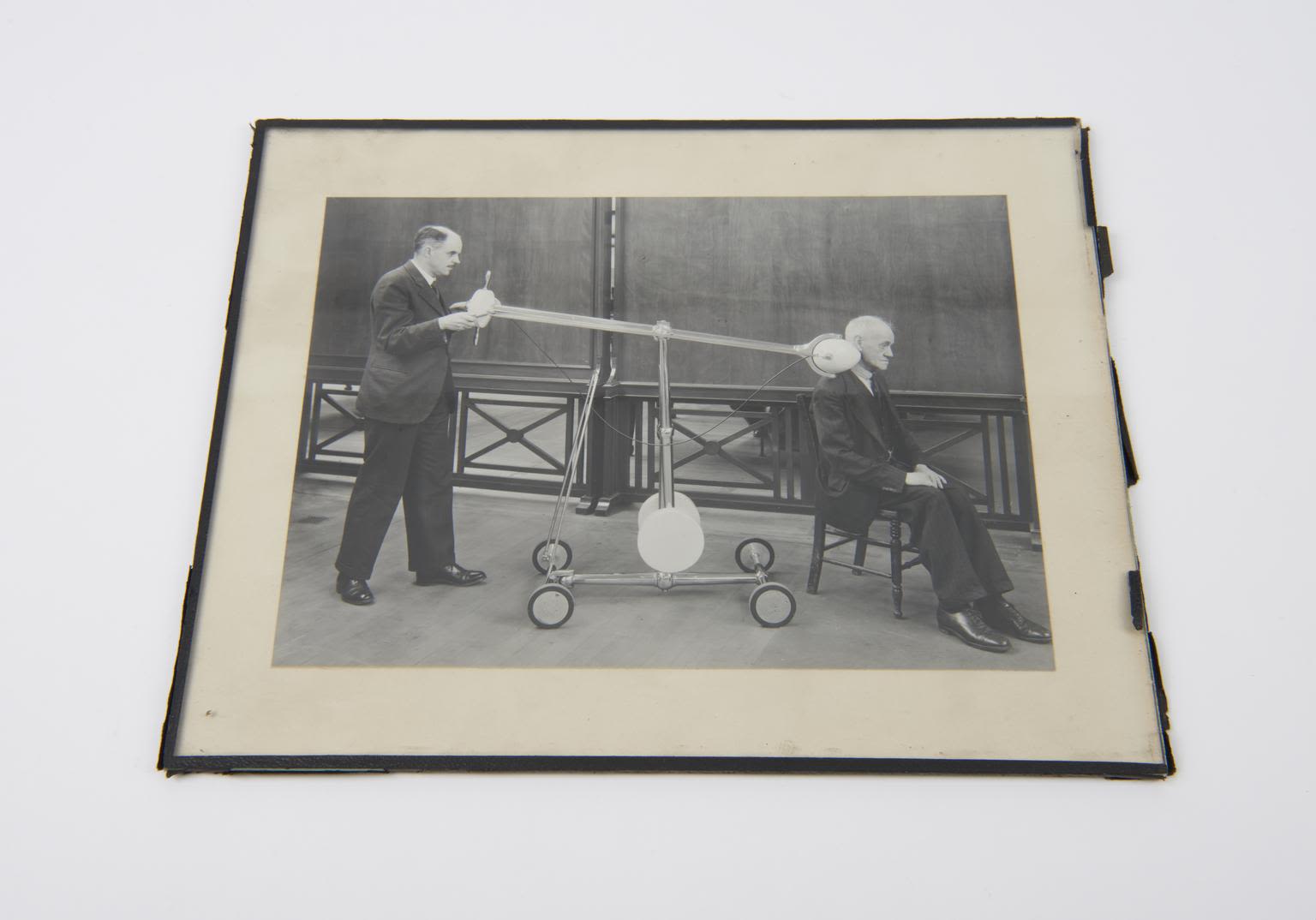 A black and white photograph showing a male doctor in a suit positioning a wheeled apparatus that looks a bit like a see-saw to deliver a radium treatment to an older male patient's neck from a distance.