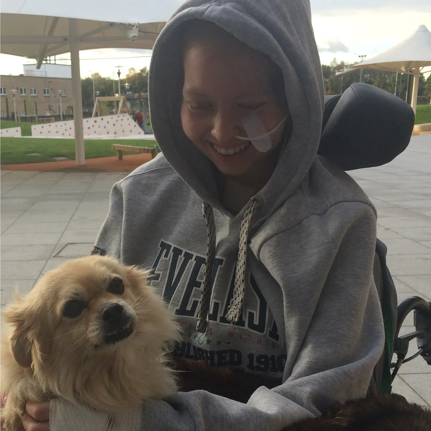 Eilidh outside in her wheelchair. She is wearing an oversized hoodie and holding a dog.