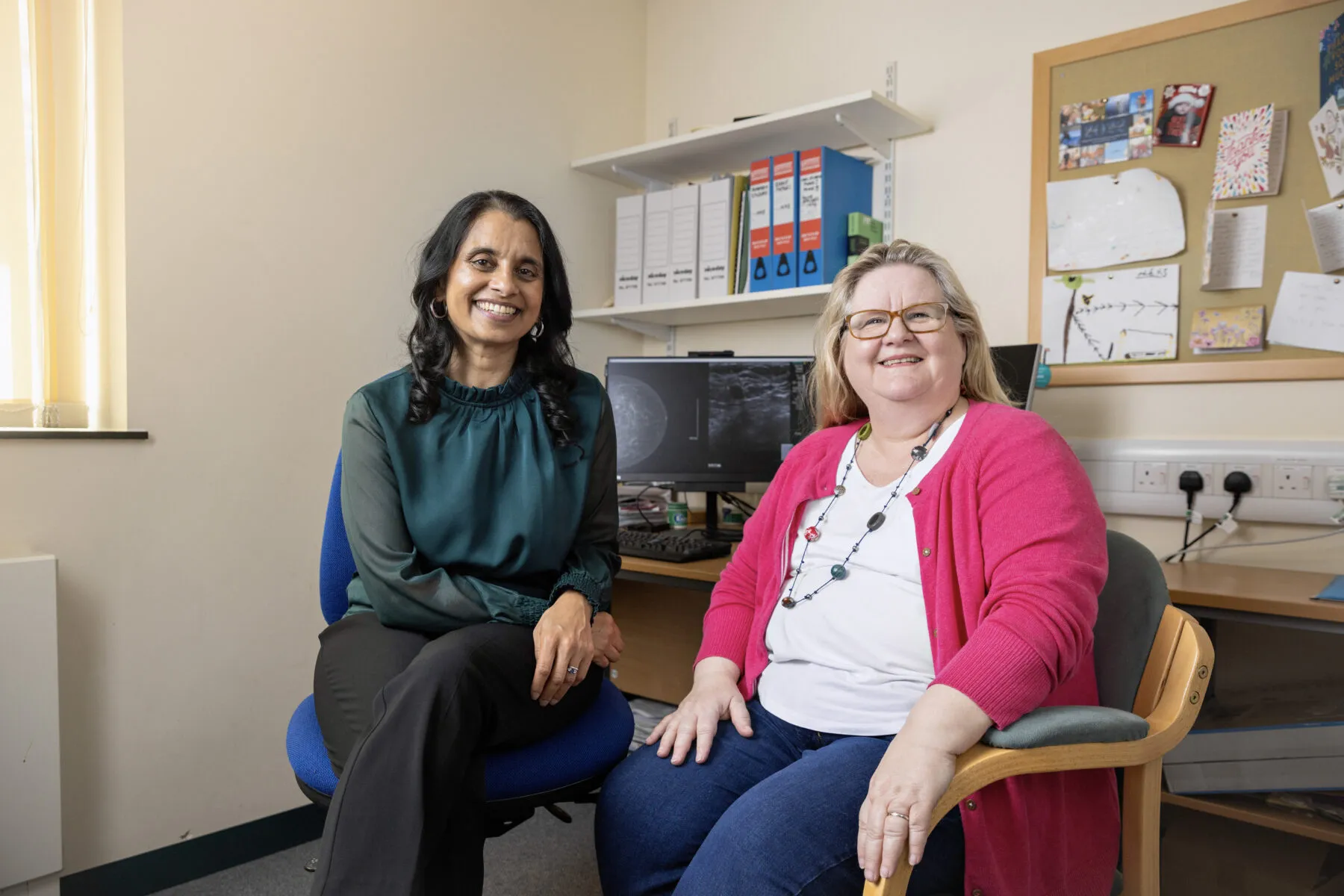 Professor Jean Abraham and PARTNER trial participant Jackie Van Bochoven together in a doctor's office. They are both looking at the camera and smiling. Behind them, a computer screen shows scans of breast tissue.
