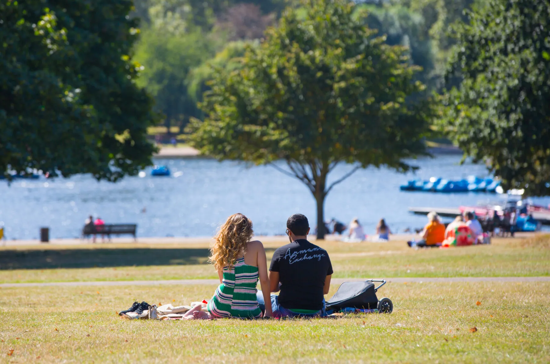 A man and woman sat in the park on a sunny day. The photograph shows them from behind, as they look out on a lake.