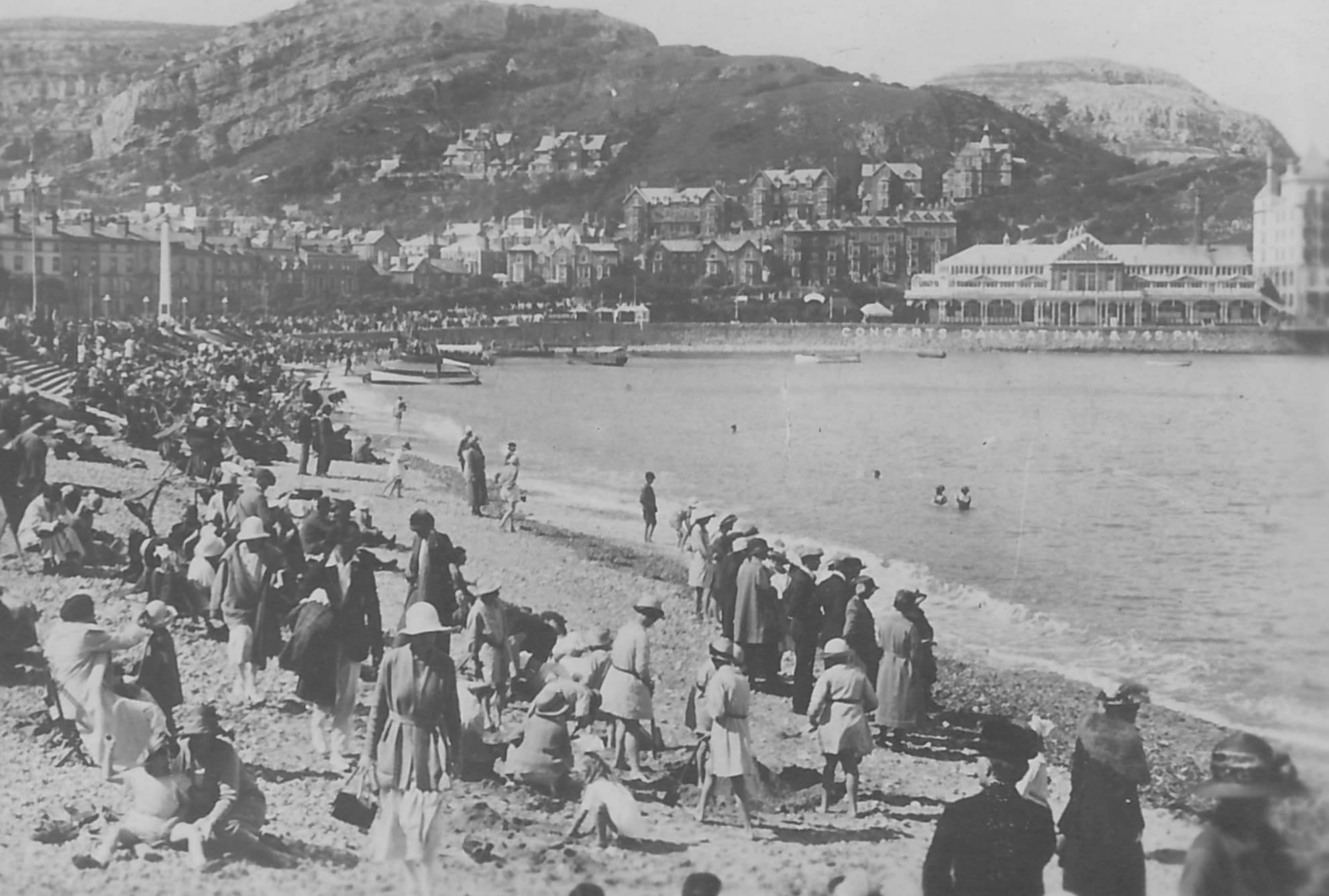 A black and white photo showing a crowd of holidaymakers on a beach. They are all wearing suits or long dresses and many have hats.  