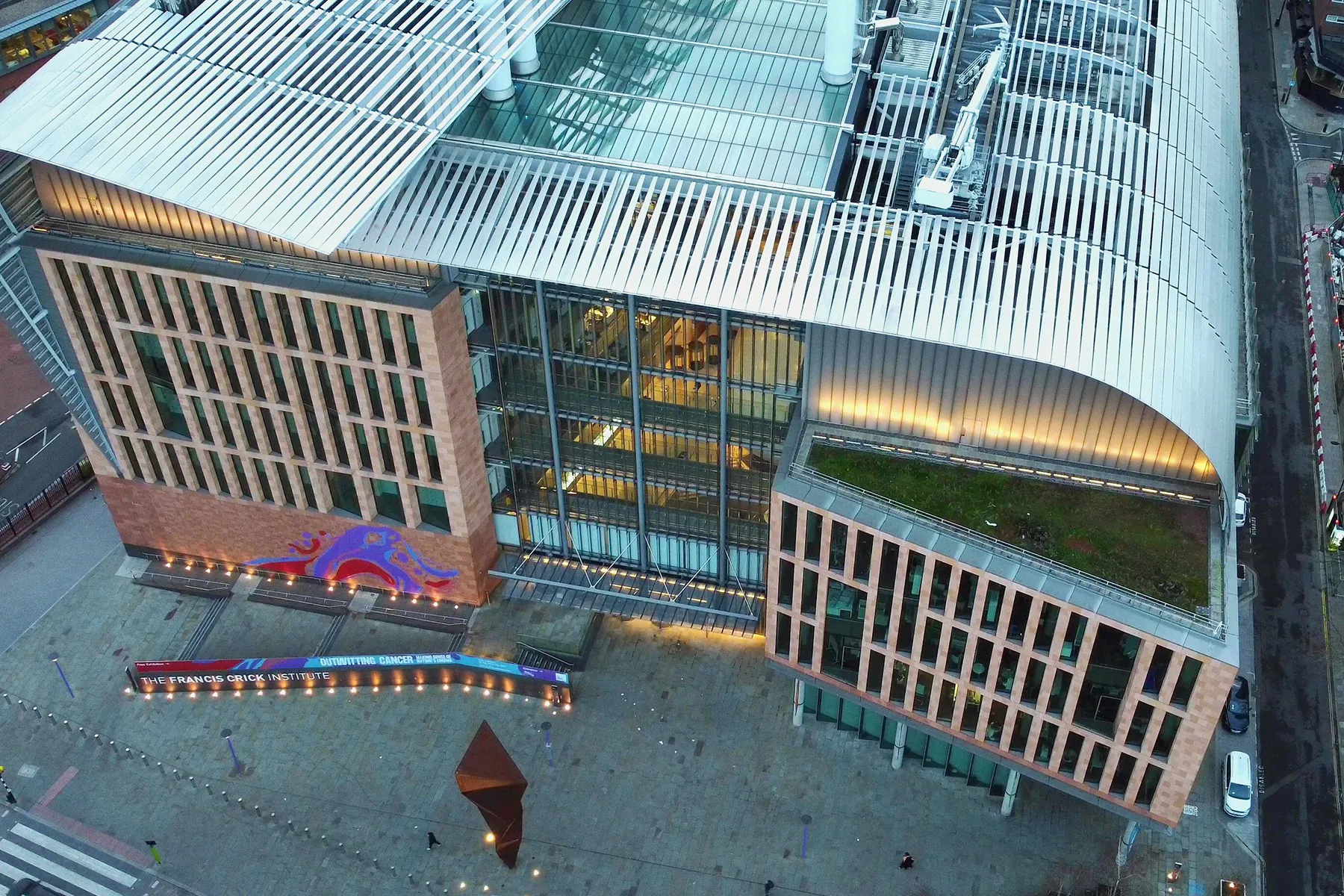 An image of the Francis Crick Institute taken by a drone. The entrance leads into a large glass central atrium, with smaller glass-panelled open-plan labs on either side.