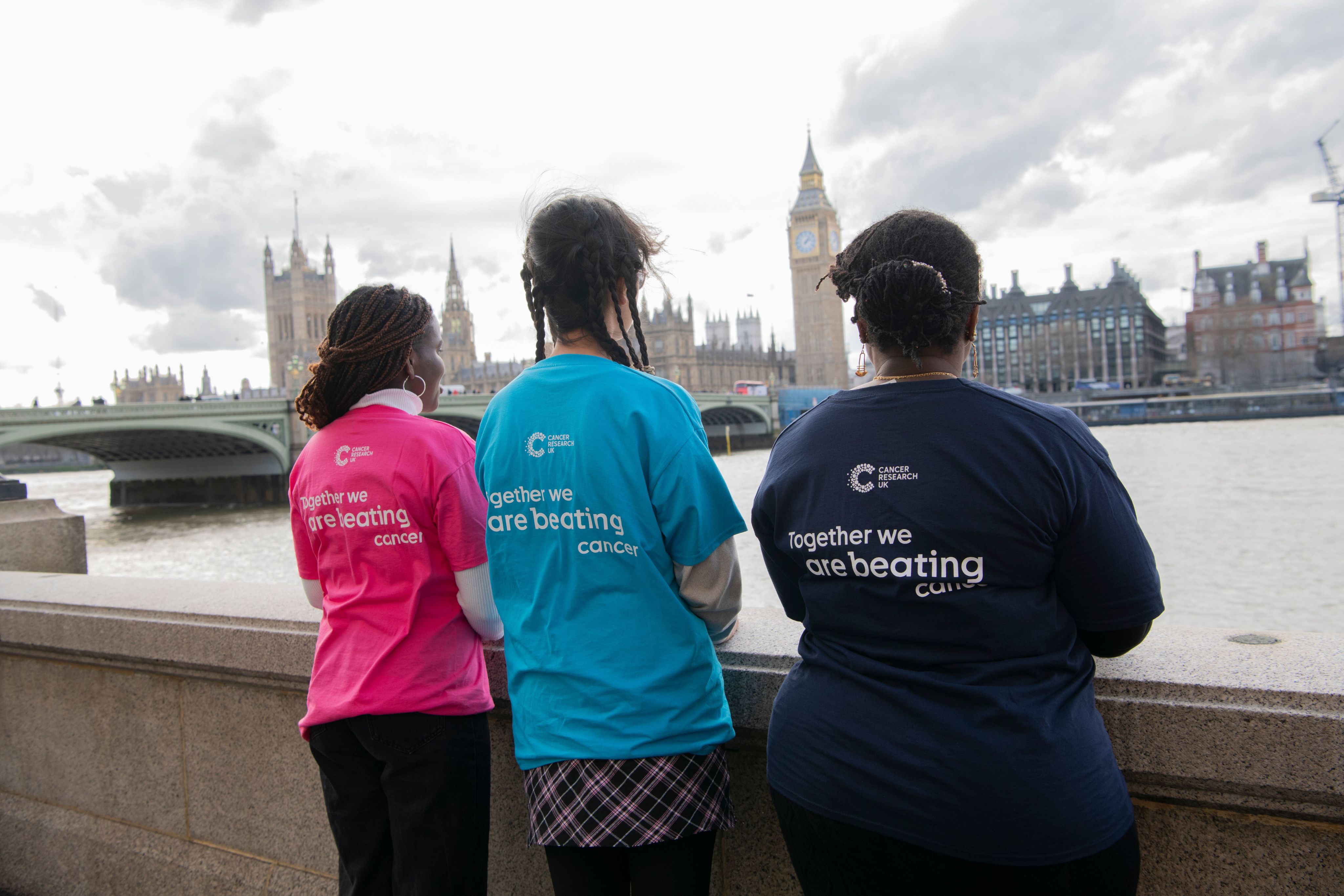 Three Cancer Research UK Campaigns ambassadors looking at the Houses of parliament in London. Their backs are to the camera and they are wearing shirts that read 'Together we are beating cancer'. 