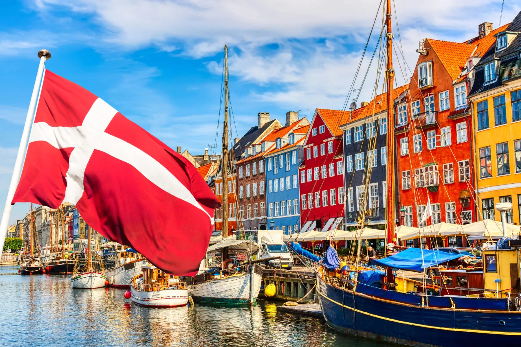 An image of the waterfront in Copenhagen. There is a Danish flag with a red background and a white cross on the left of the picture. In the background there are colourful buildings and sailboats.