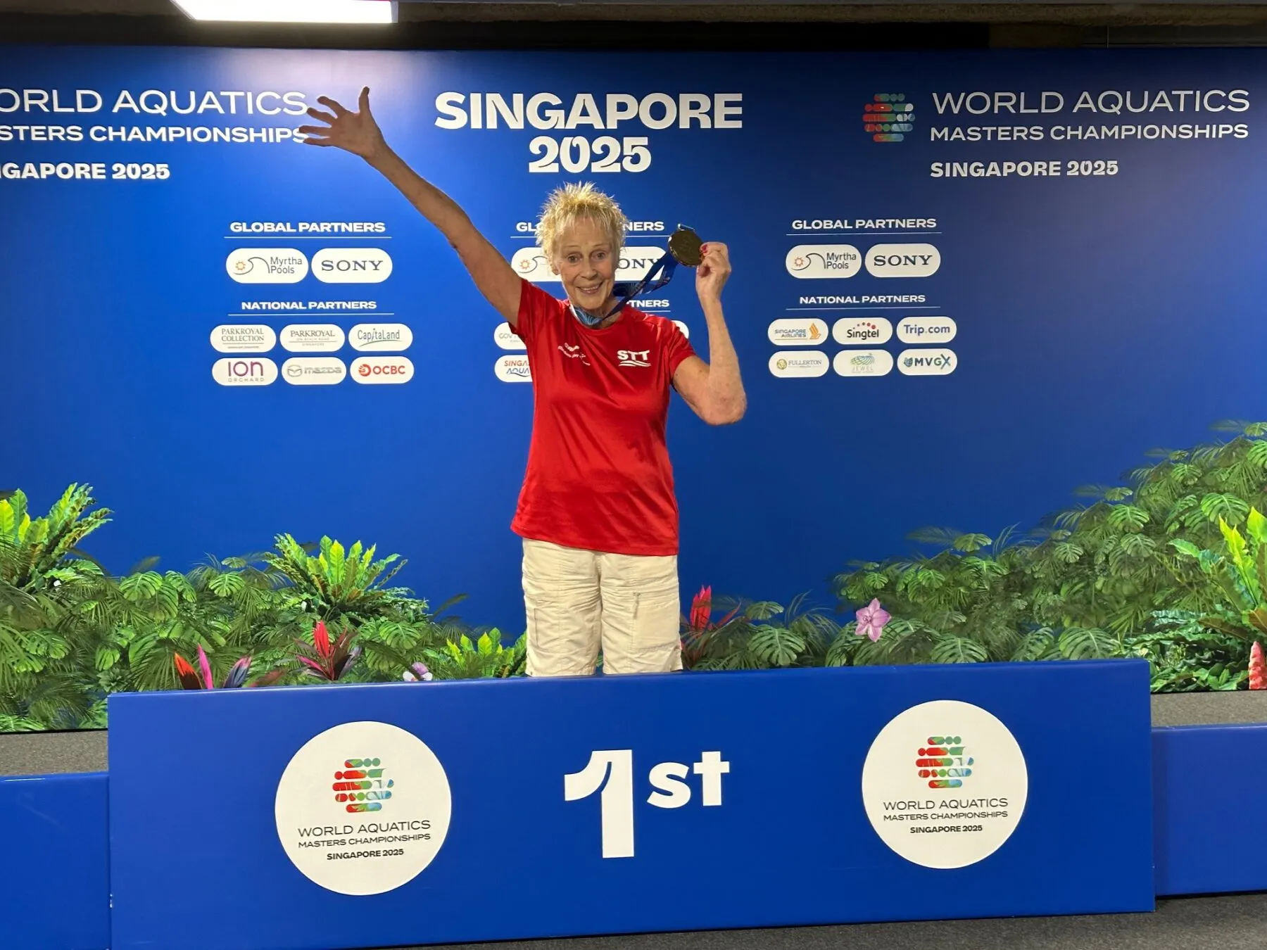 Elisabeth stands on a blue winners podium in Singapore, holding her medal after winning a world swimming competition. She is wearing a red shirt and white shorts.