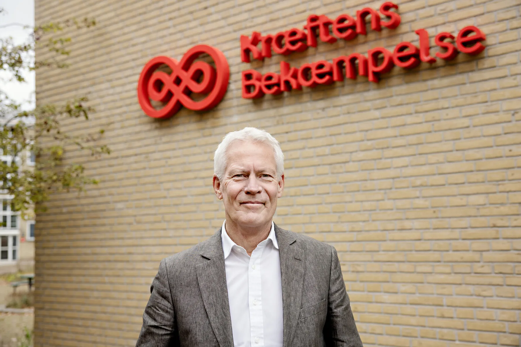 A professional headshot of Jesper Fisker, dressed in a grey blazer and white shirt, standing outside the headquarters of the Danish Cancer Society.