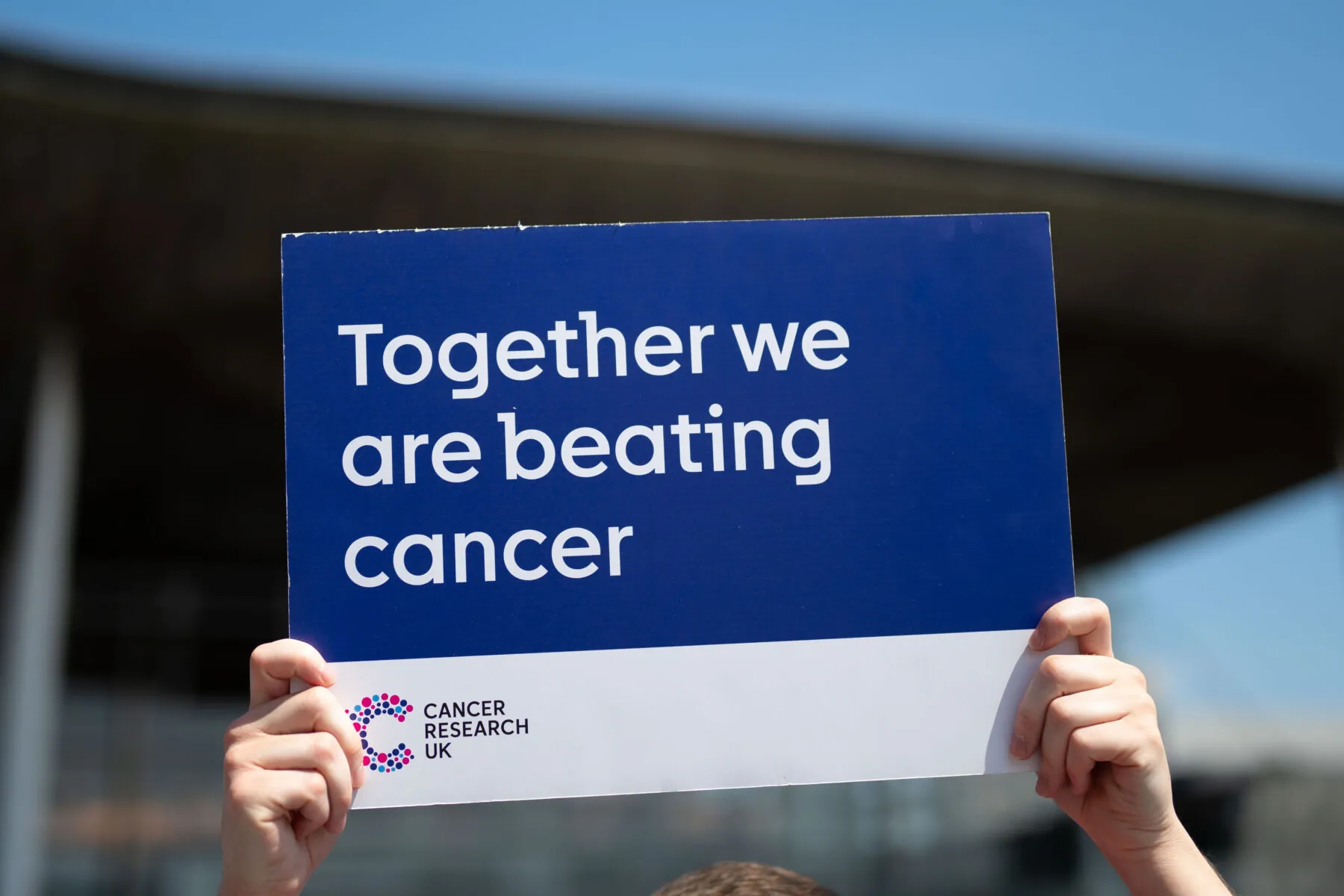 Two hands holding up a blue placard that says 'Together we are beating cancer' in white text. There is a Cancer Research UK logo in the bottom left of the placard. The Welsh Senedd building is visible in the background. It is a sunny day with a clear blue sky.