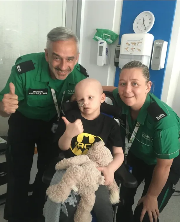 Jack sat in his wheelchair holding his teddy. On his left and right are two St John's Ambulance drivers, Paul and Jackie.