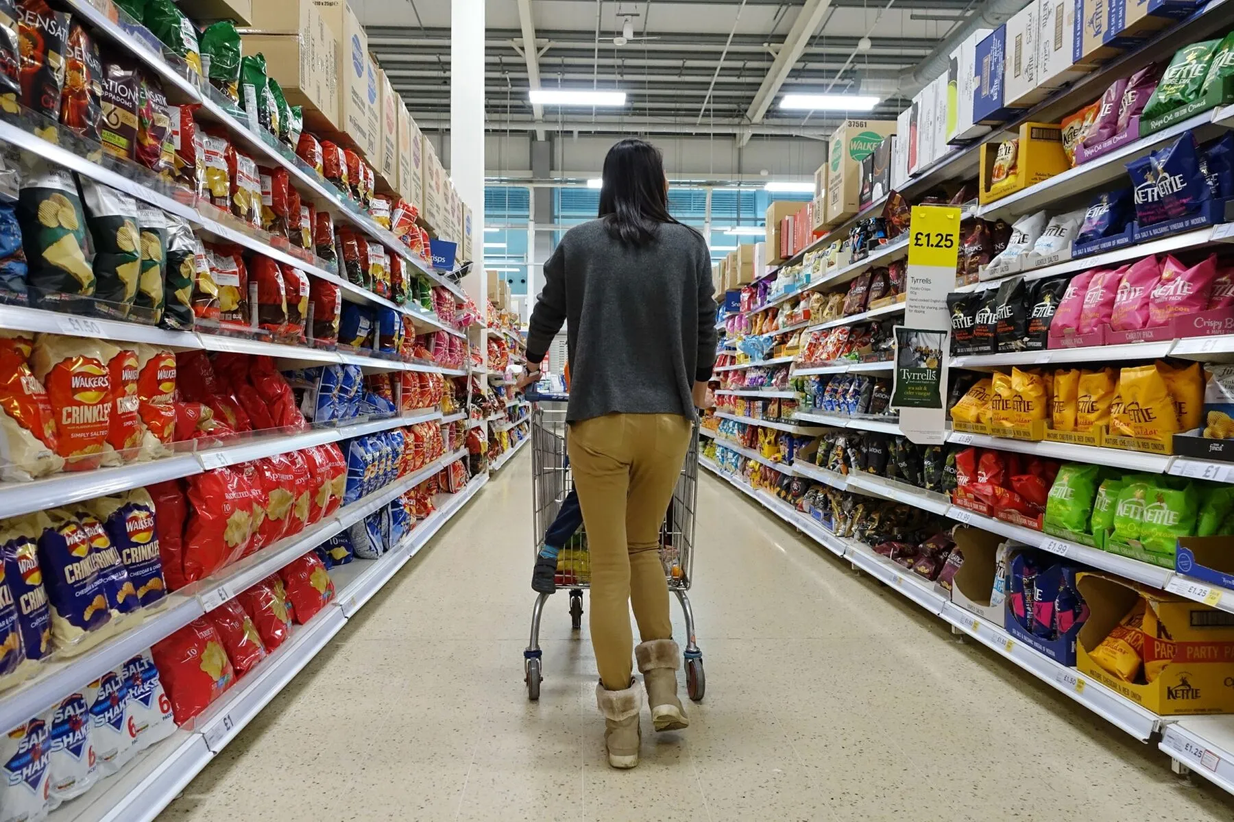 A shopper pushes a trolley down a supermarket aisle full of brightly coloured crisp packets.