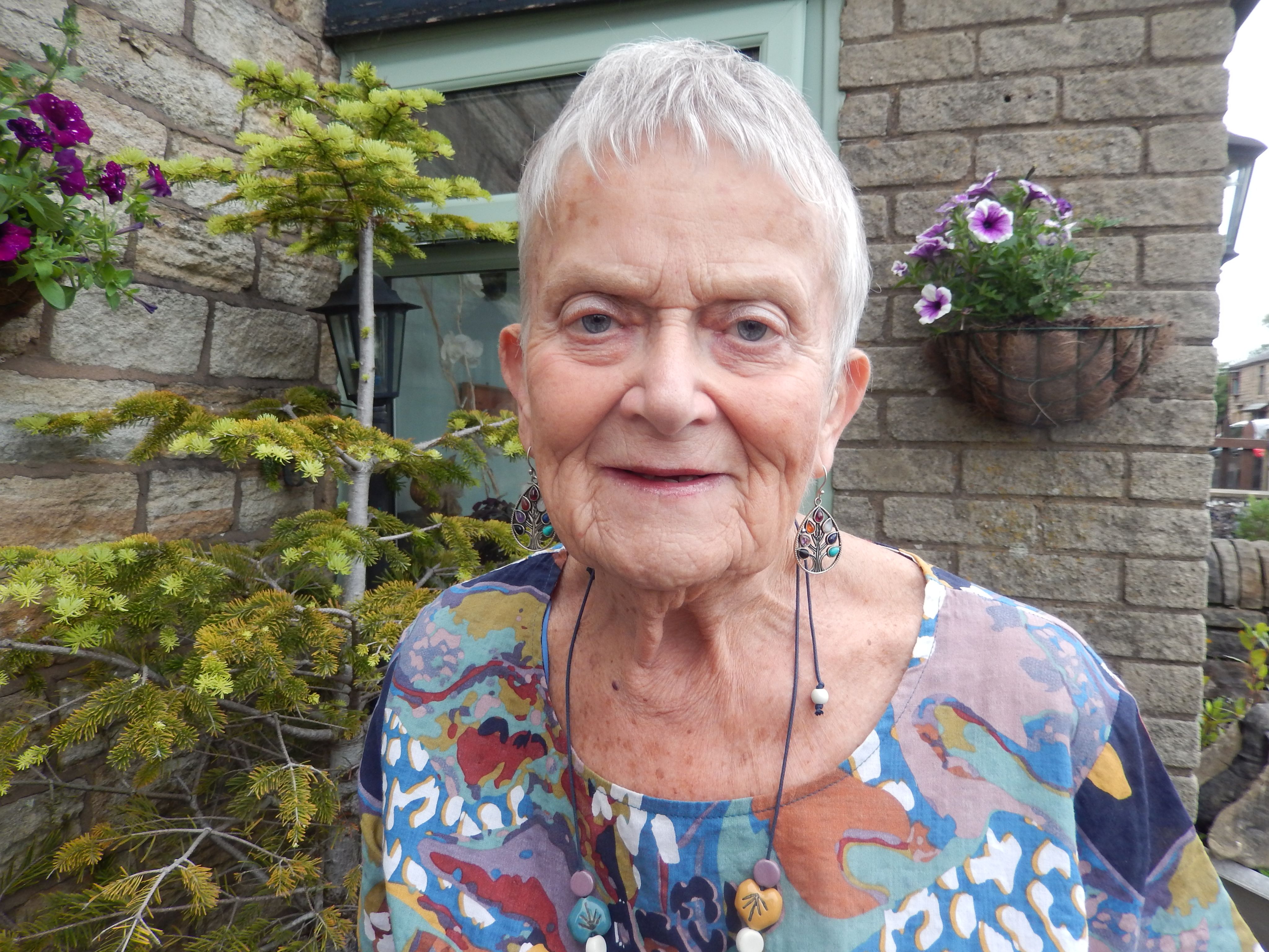 Carol outside her home with a tree and hanging baskets filled with purple flowers.