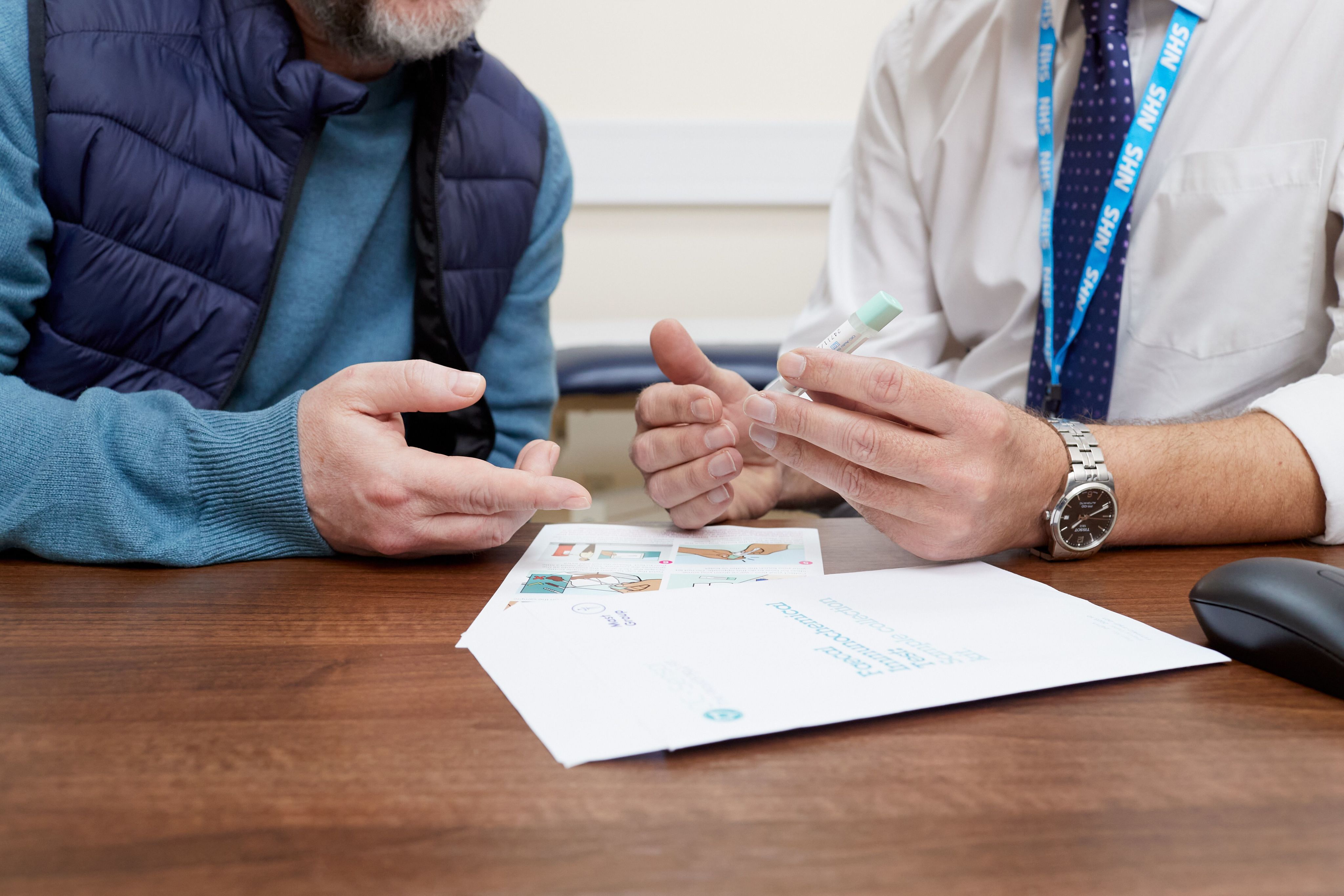 A GP showing a bowel screening (FIT) test to a patient. They are sat at the desk in the GP's office. The camera is focused on their hands and their faces aren't visible.