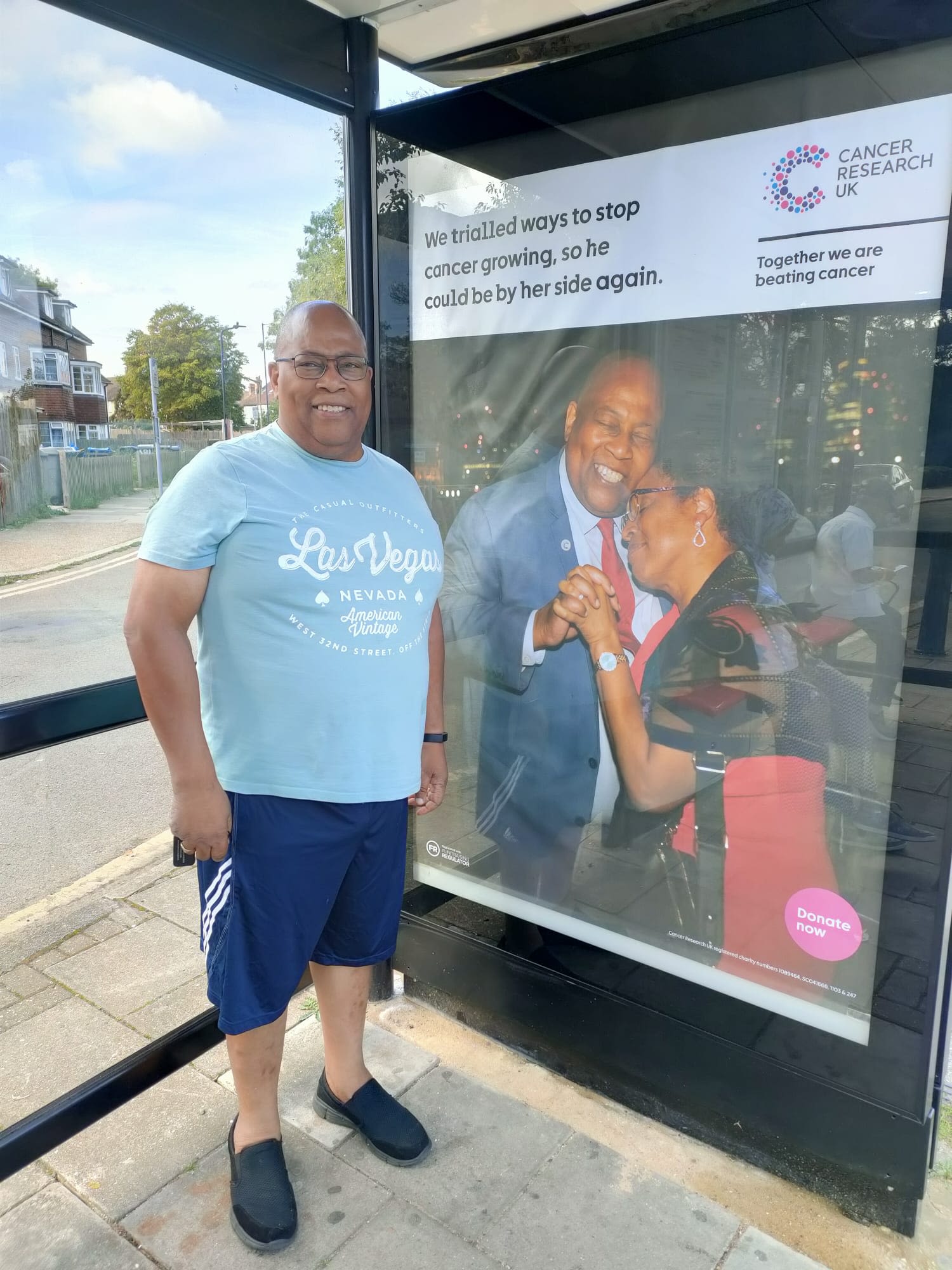 Alfred next to a Cancer Research UK poster which shows him and his wife embracing. The poster reads: 'We trialled ways to stop cancer growing, so he could be by her side again.'