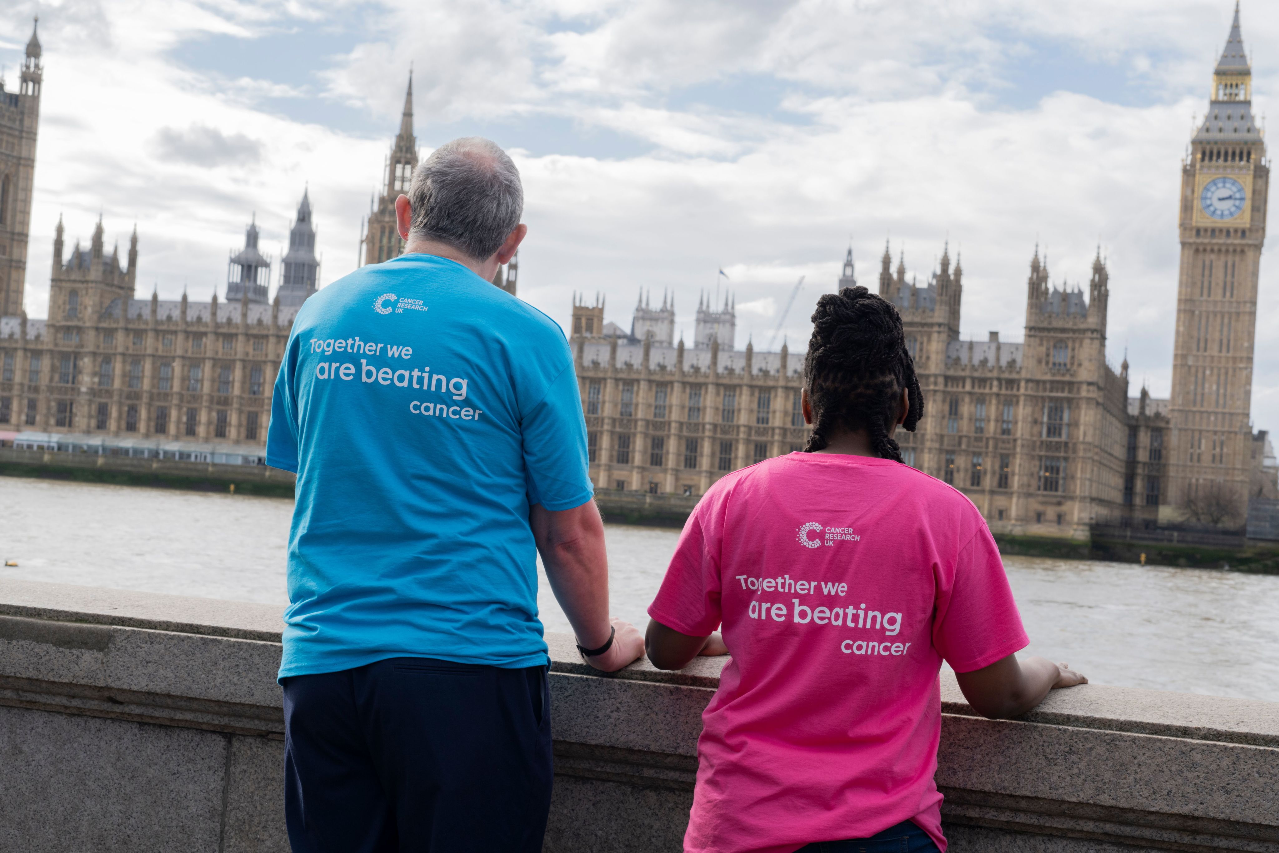 Two Cancer Research UK Campaigns ambassadors looking at the Houses of parliament in London. Their backs are to the camera and they are wearing shirts that read 'Together we are beating cancer'. 