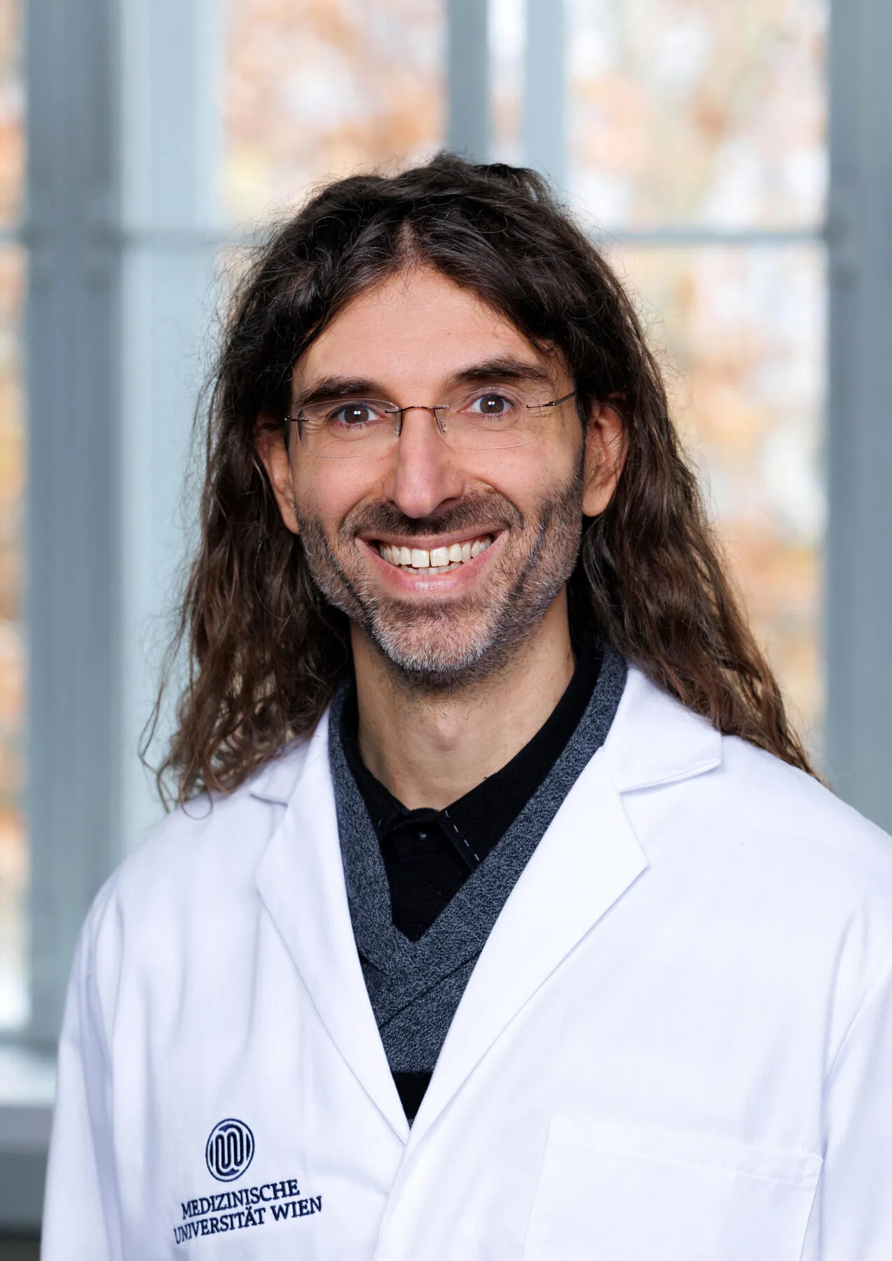 Dr Saverio Tardito, a long-haired, bearded man with a broad smile, in his new lab in Vienna, He has small rimless glasses over his eyes and is wearing a lab coat over a black shirt and grey jumper.