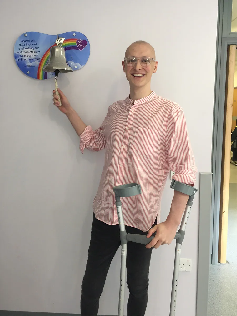 Cory ringing a bell in hospital after finishing one of his courses of chemotherapy. He's smiling widely in glasses, a striped pink shirt and black jeans. He's ringing the bell with his right hand while supporting two crutches in his left.