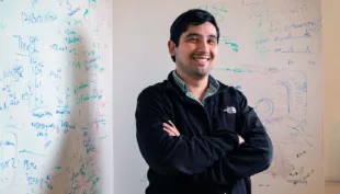 Dr Jason Buenrostro, Harvard University professor, smiling in a black with arms crossed in front of whiteboards with math equations written in green and blue