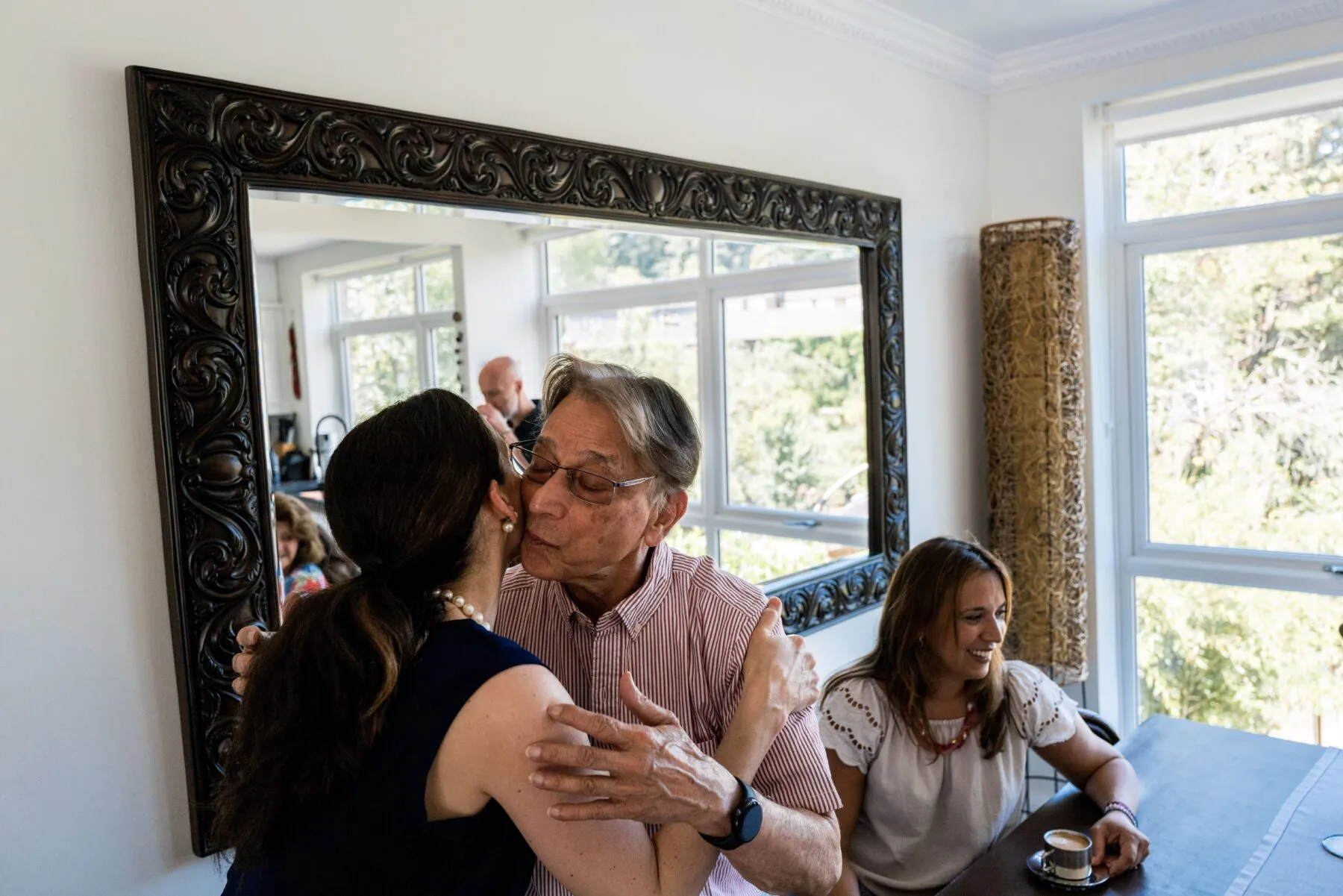 A family gathering for lunch. In the foreground, two people are kissing each other's cheeks in greeting.
