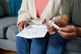 A man with his partner on the sofa opening a bowel cancer screening test kit from NHS England.