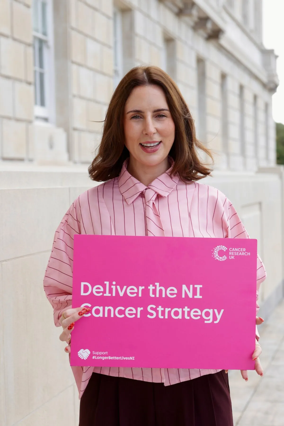 Women holding a pink sign that says 'Deliver the NI Cancer Strategy'
