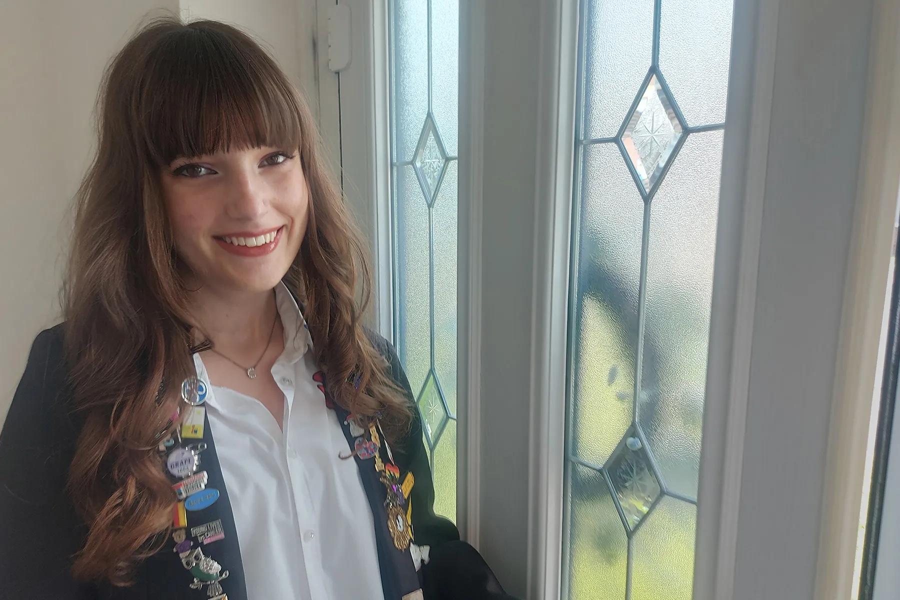 A teenage girl in her school uniform smiling as she opens her front door to go out. She has lots of badges on the lapels of he school blazer.