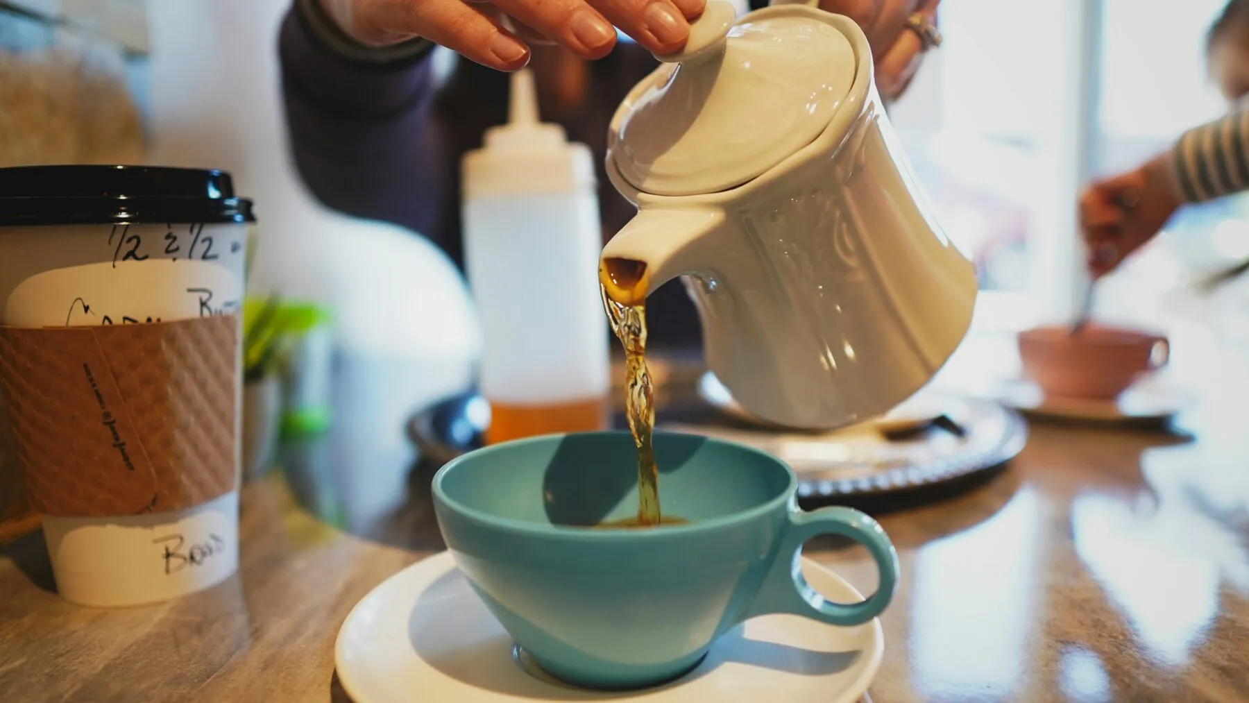 Close up of someone pouring tea from a white teapot into a blue teacup on a white saucer.
