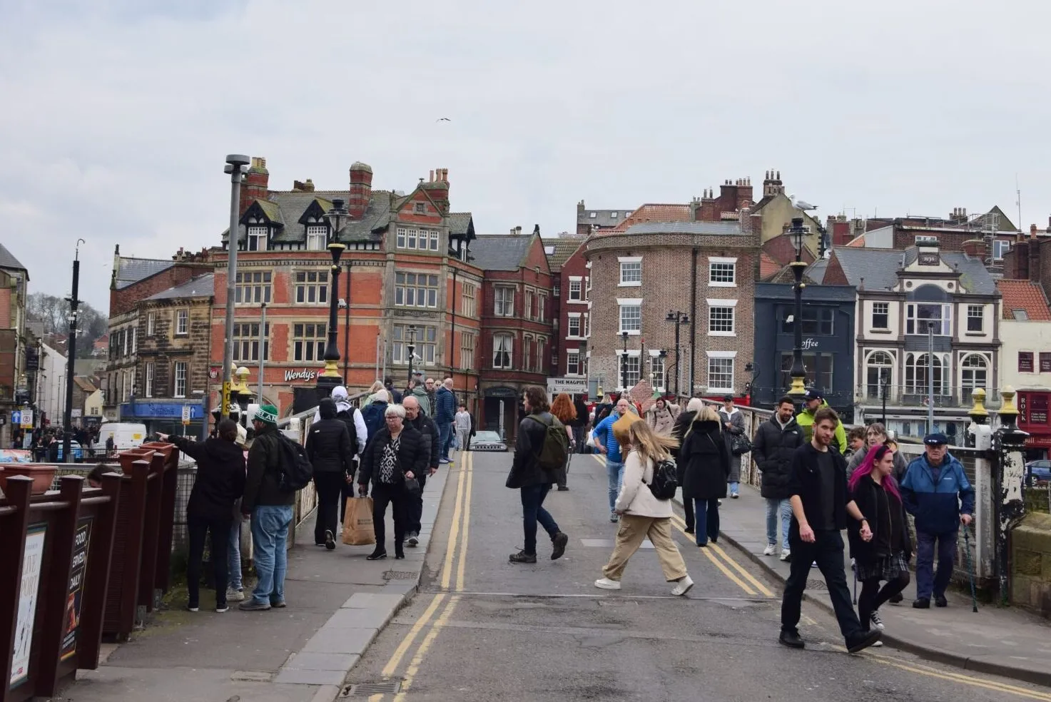 People crossing road with buildings visible in the background against a cloudy sky.
