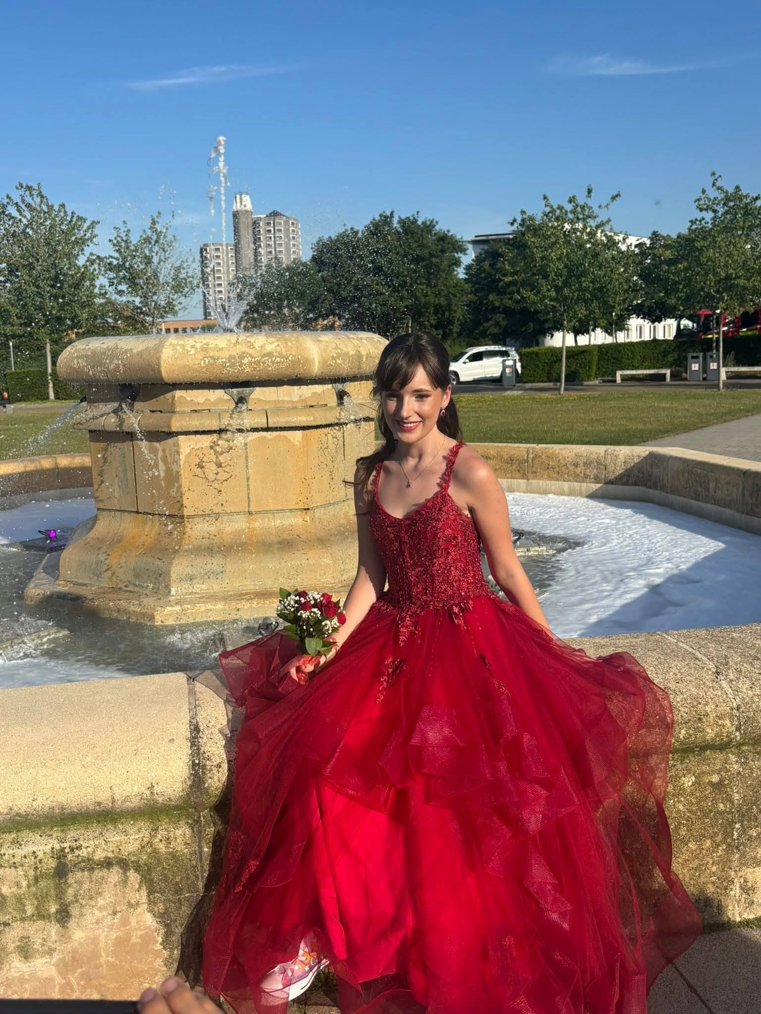 Caitlin in her red prom dress leaning on the wall of a fountain, holding a bouquet of flowers.