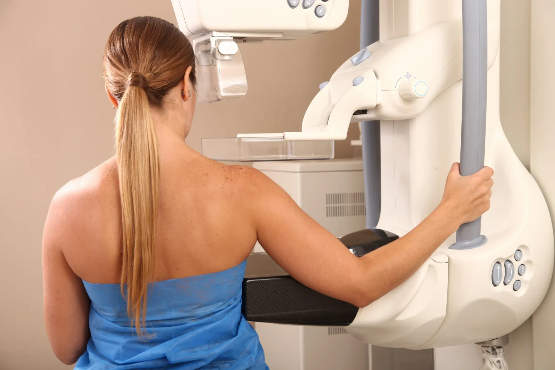 A woman seen from behind preparing for a mammogram. She has long blonde hair tied in a pony tail and is holding a handle on the mammography machine in front of her.
